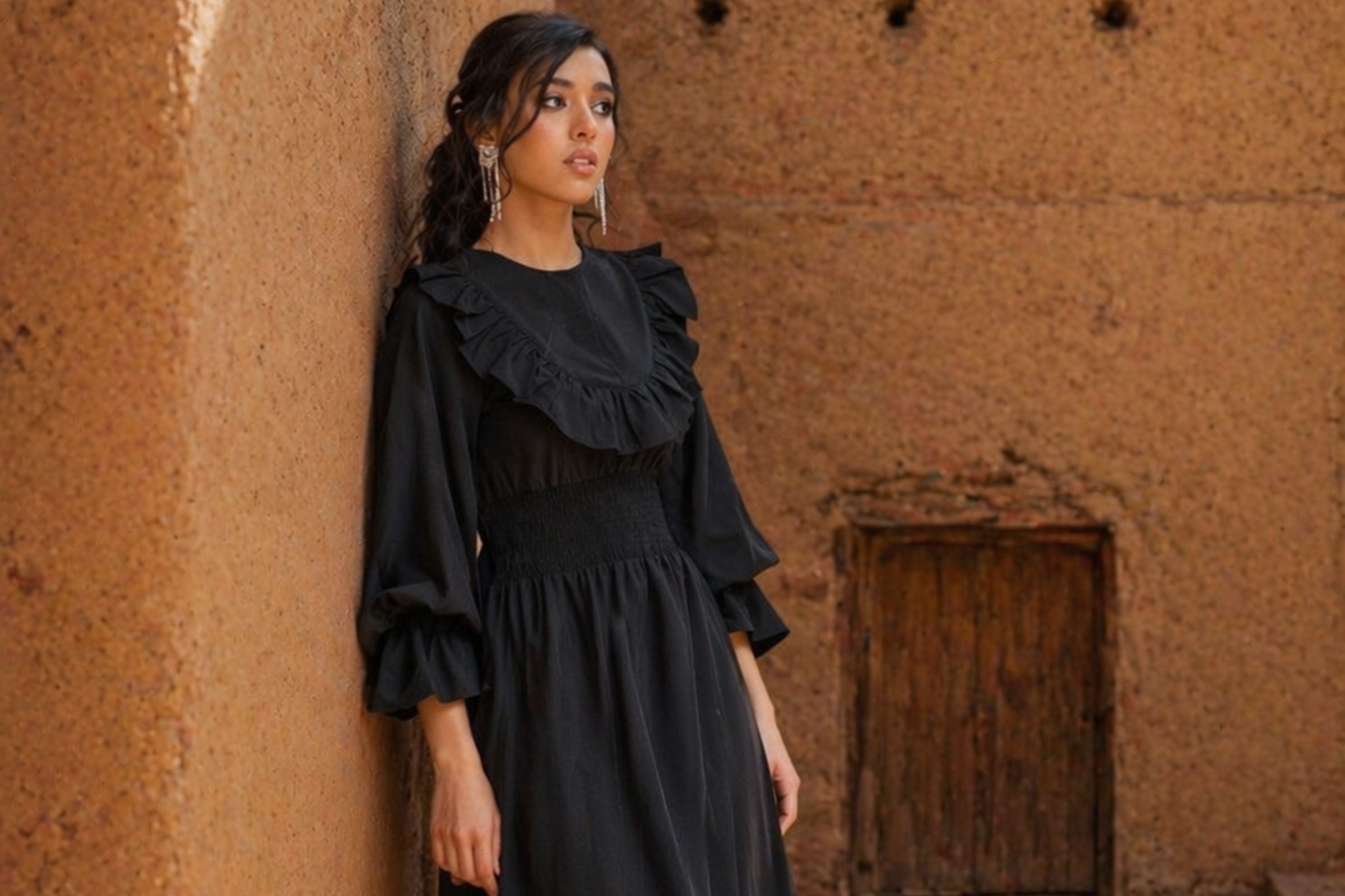 A woman wearing a black long-sleeve dress with dramatic ruffles and a smocked waist, standing against a warm, textured adobe wall.