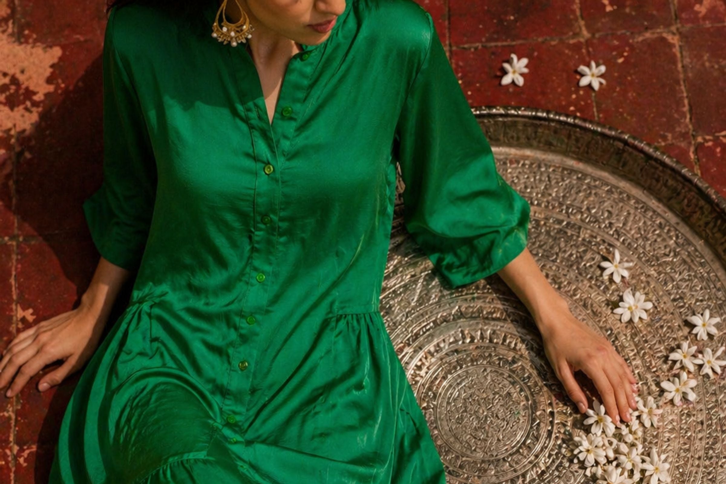 A woman wearing a shiny emerald green satin button-up dress sits on a round patterned tray against a reddish-brown tiled background.