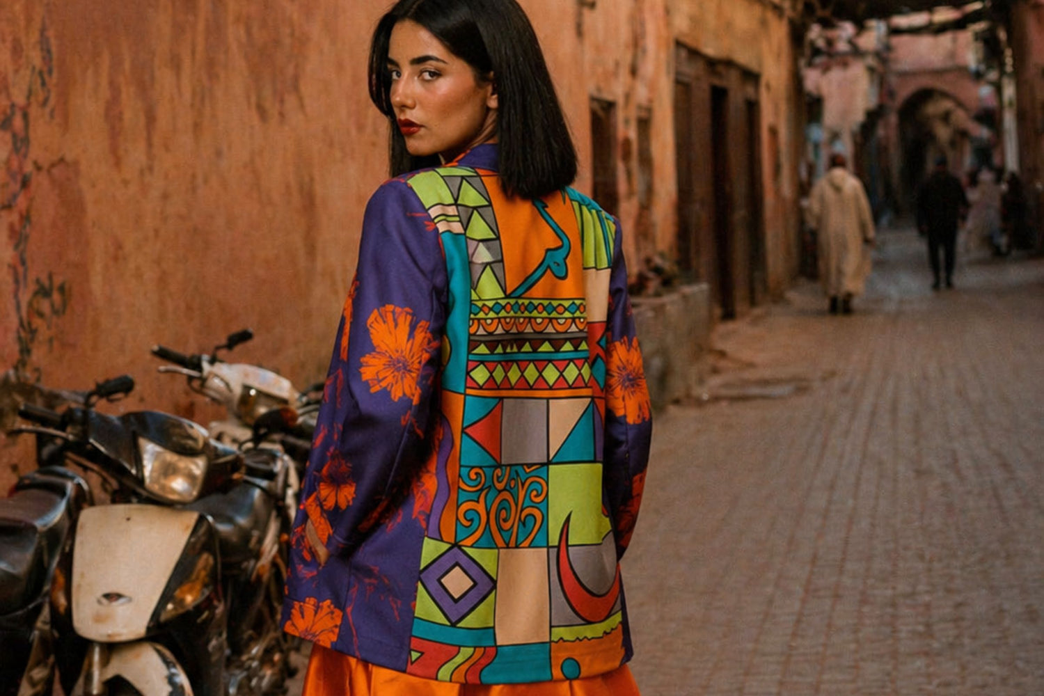 A woman stands in a narrow outdoor street wearing a colorful patterned jacket with geometric and floral motifs in purple, orange, green, yellow, and blue. Motorbikes are parked on the left, and buildings line the background.