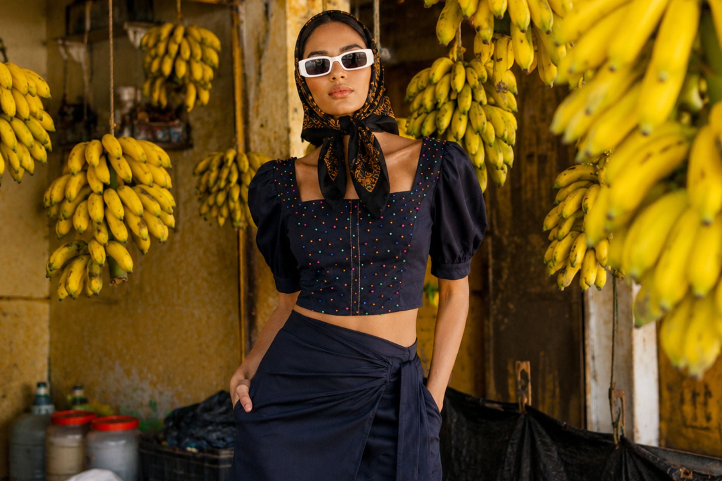 A woman wearing a dark navy blue cropped puff-sleeve top with colorful polka dots, a matching dark skirt, white sunglasses, and a patterned headscarf stands in a market setting surrounded by hanging yellow bananas.