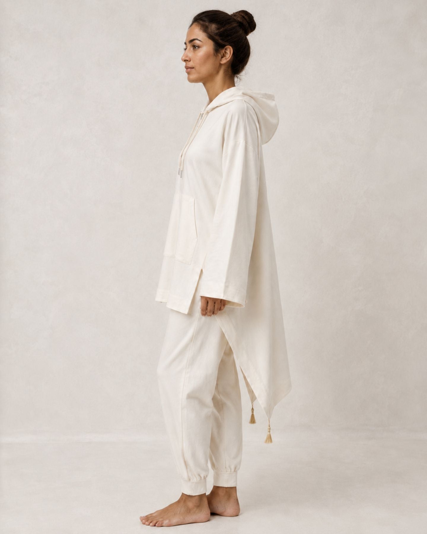 A woman stands in profile wearing a loose, long-sleeve cream-colored hooded robe or poncho with matching jogger pants, photographed against a plain light beige studio backdrop.