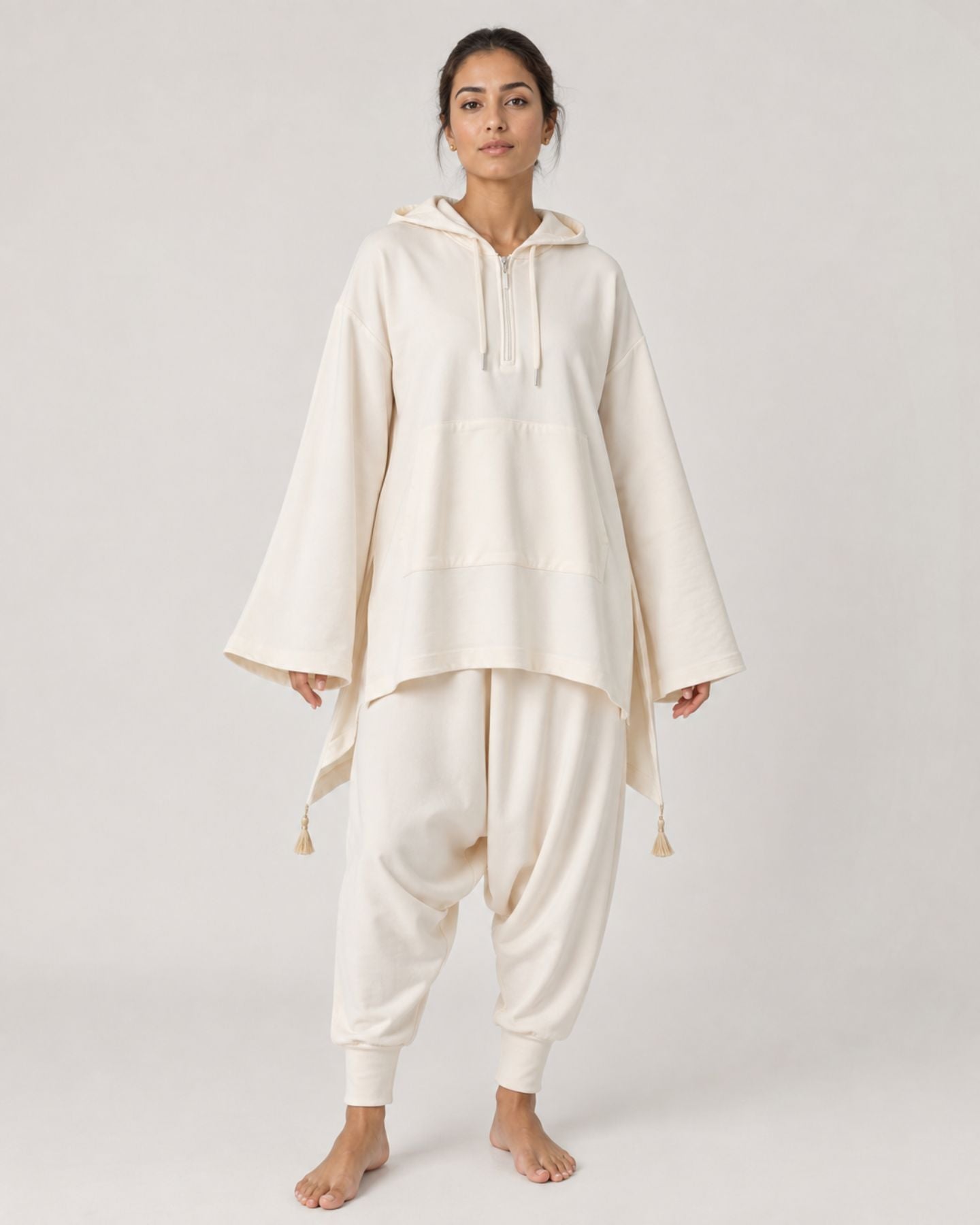A woman modeling a cream-colored hooded tunic and matching baggy pants against a plain light gray studio background.